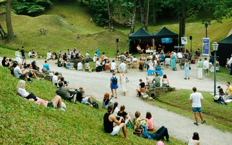 Pulcēšanās brīvā dabā parkā, kur cilvēki sēž uz zāles un krēsliem, vērsti pret melnu telti. Noskaņojums ir mierīgs sulīgi zaļojošu augu vidū.