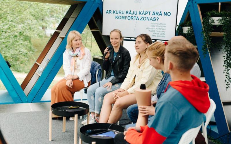 Seši cilvēki sēž puslokā ārā un iesaistās dzīvā diskusijā. Sieviete smaida un tur mikrofonu. Fonā ir uzraksts "Ar kuru kāju kāpt ārā no komforta zonas?".
