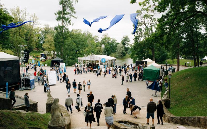 Brīvdabas festivāla aina ar cilvēku pūli, kas pulcējas koku ieskautā laukumā. Virs galvas plīvo zili karogi. Redzamas teltis un skatuve, kas liecina par dzīvīgu, svētku atmosfēru.