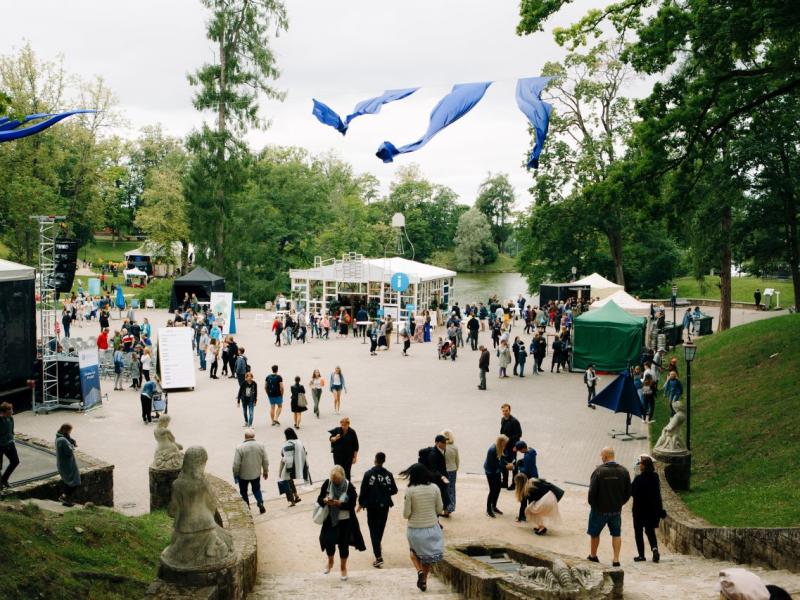 Brīvdabas festivāla aina ar cilvēku pūli, kas pulcējas koku ieskautā laukumā. Virs galvas plīvo zili karogi. Redzamas teltis un skatuve, kas liecina par dzīvīgu, svētku atmosfēru.