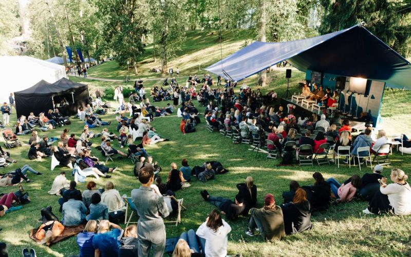 Festivāla aina brīvā dabā parkā ar zilu telti, kurā notiek paneļdiskusija. Dažāda auditorija sēdvietās, relaksēta atmosfēra koku ieskautā.