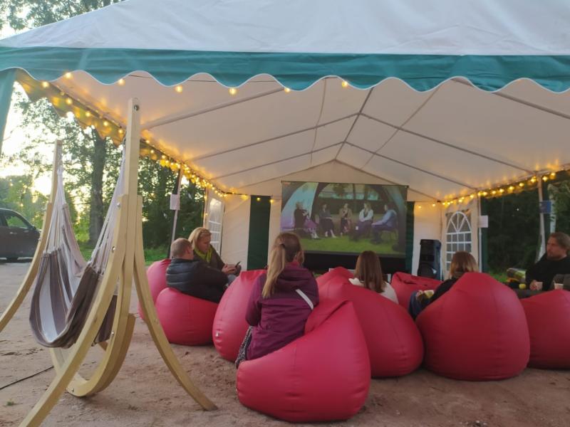 Mājīga brīvdabas filmu seanss zem telts ar virtenēm. Cilvēki atpūšas sarkanos pupu maisos, skatoties pret ekrānu, radot siltu, kopīgu atmosfēru.