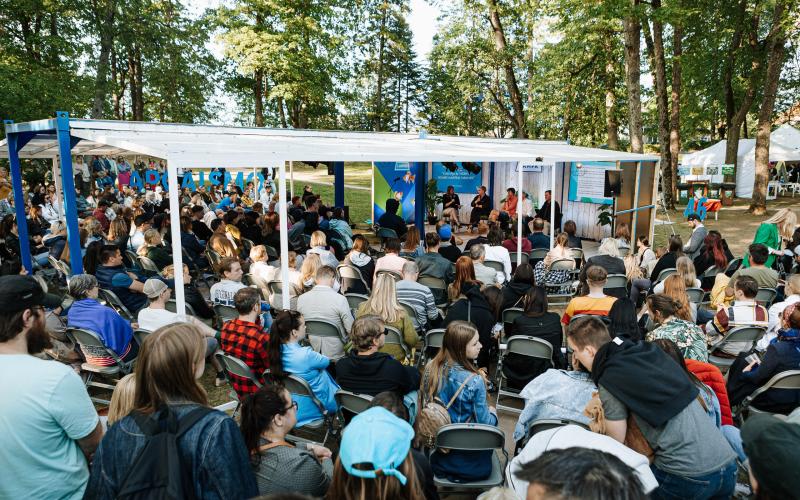 Āra pasākums ar lielu pūli, kas sēž zem baltas nojumes, ko ieskauj koki. Cilvēki ir uzmanīgi, baudot dzīvīgu diskusiju mierīgā gaisotnē.