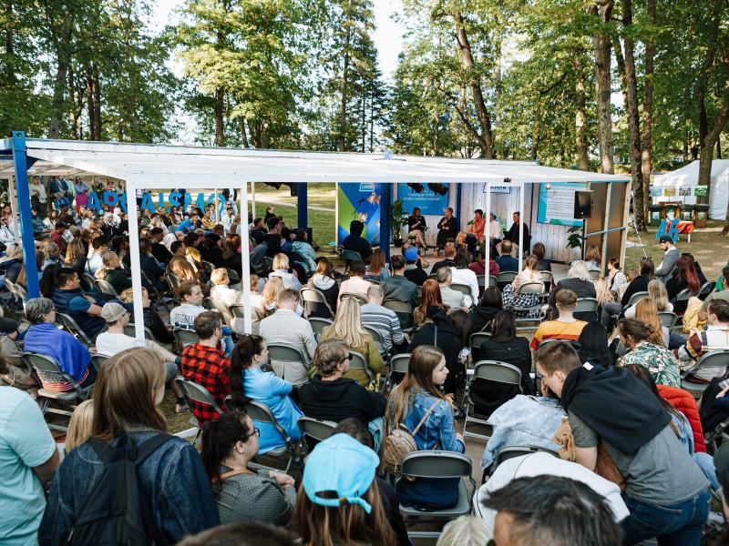 Āra pasākums ar lielu pūli, kas sēž zem baltas nojumes, ko ieskauj koki. Cilvēki ir uzmanīgi, baudot dzīvīgu diskusiju mierīgā gaisotnē.