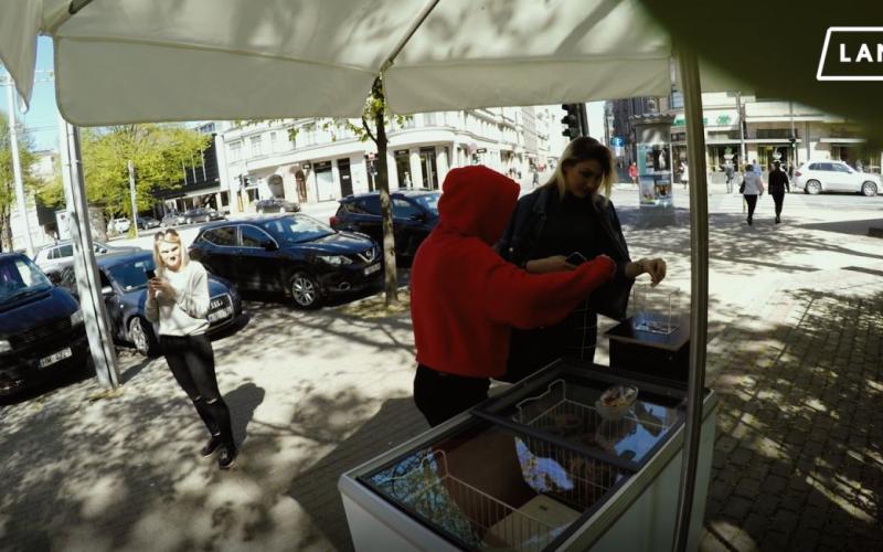 Saulaina ielasaina, kurā cilvēks sarkanā kapučjakā veic darījumu pie pārdevēja ratiņiem. Netālu pastaigājas sieviete, turot telefonu, un viņu ieskauj novietotas automašīnas un koki, radot dzīvīgu pilsētvides atmosfēru.