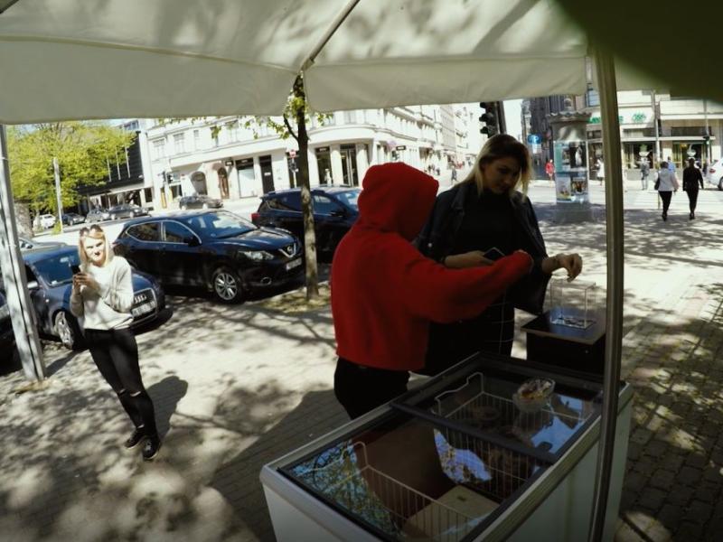 Saulaina ielasaina, kurā cilvēks sarkanā kapučjakā veic darījumu pie pārdevēja ratiņiem. Netālu pastaigājas sieviete, turot telefonu, un viņu ieskauj novietotas automašīnas un koki, radot dzīvīgu pilsētvides atmosfēru.