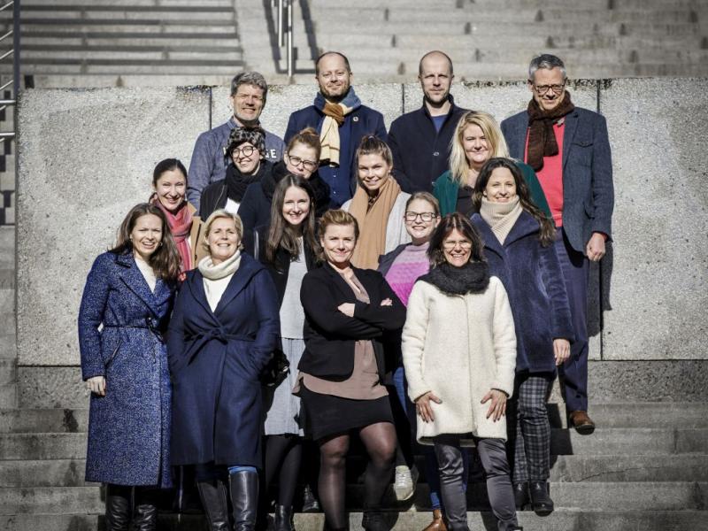 Sešpadsmit cilvēku grupa stāv uz akmens pakāpieniem spožā saules gaismā, smaidoši un ziemas drēbēs. Noskaņojums ir priecīgs un sadarbīgs.