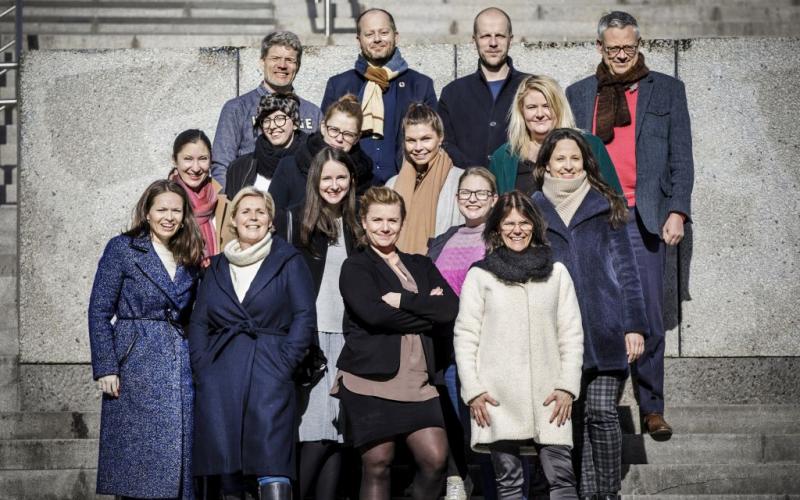 Sešpadsmit cilvēku grupa stāv uz akmens pakāpieniem spožā saules gaismā, smaidoši un ziemas drēbēs. Noskaņojums ir priecīgs un sadarbīgs.