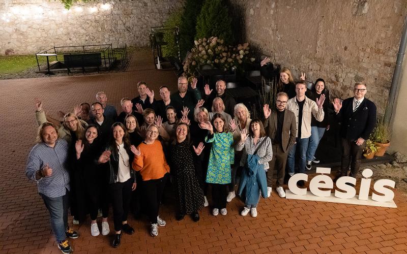 Smaidīgu cilvēku grupa māj kamerai ārā uz ķieģeļu terases, uz zemes lieliem burtiem rakstīts "Cēsis".