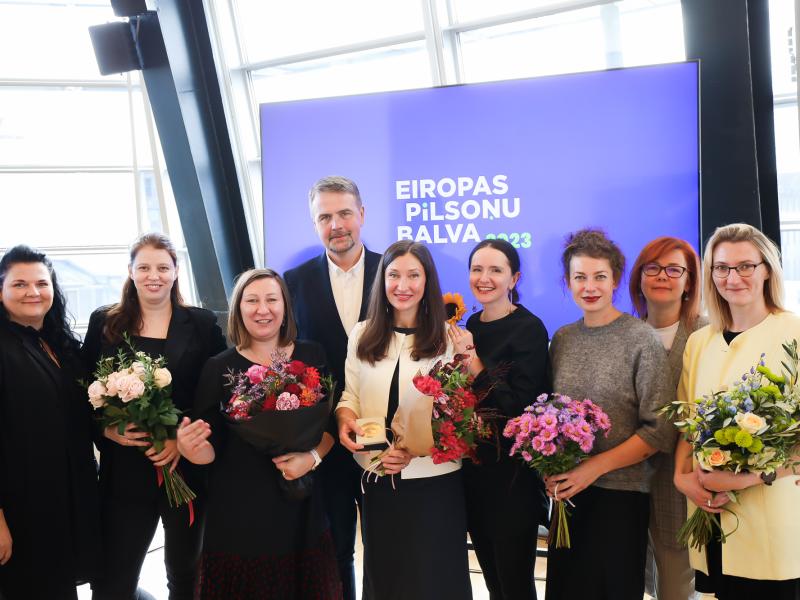 Deviņu cilvēku grupa smaida iekštelpās, rokās turot ziedus. Fonā esoša zīme ar uzrakstu "Eiropas Pilsoņu Balva 2023", kas norāda uz apbalvošanas pasākumu.