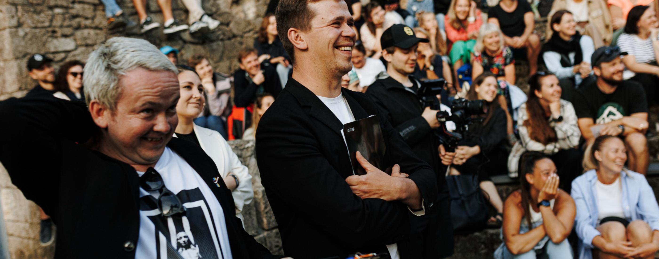 Dažādu cilvēku grupa, tostarp divi vīrieši - Edgars Bāliņš un Rūdolfs Kugrēns -priekšplānā smejoties, sēž ārā uz akmens pakāpieniem. Noskaņojums ir priecīgs un mierīgs, daži cilvēki iemūžina mirkļus ar kamerām.