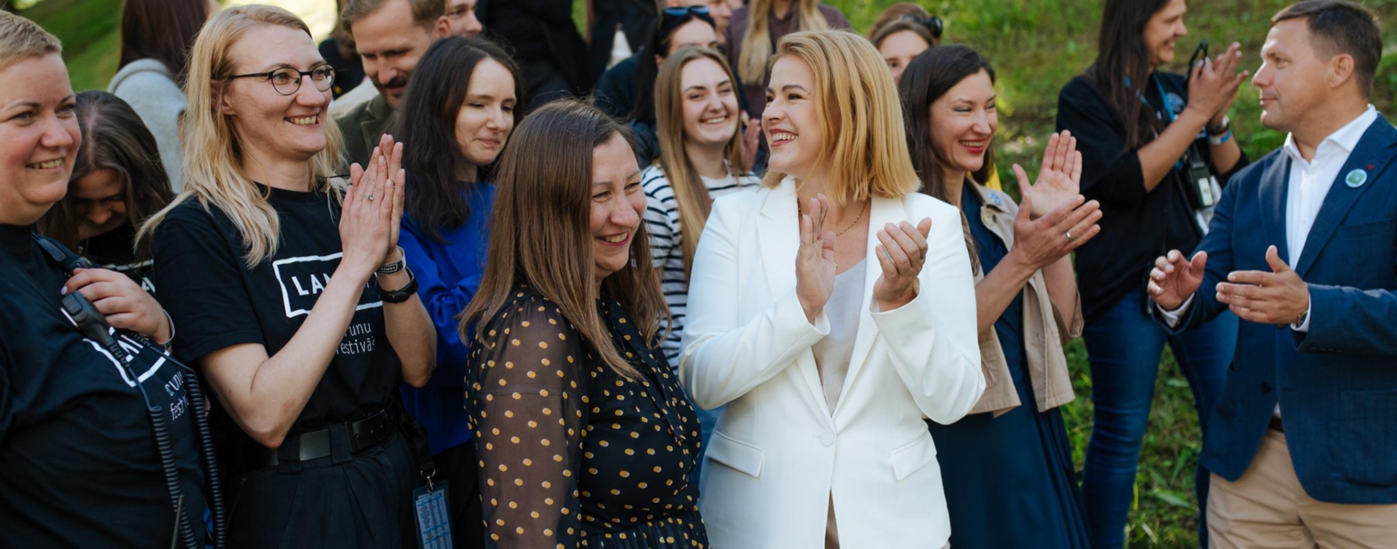 Smaidīgu cilvēku grupa ārā, aplaudējot un izbaudot jautru mirkli. Priekšplānā Latvijas Ministru prezidente Evika Siliņa, Sarunu festivāls LAMPA direktore Egita Prāma. Aina ir dzīvīga un silta, ar saules gaismu un zaļu zāli.