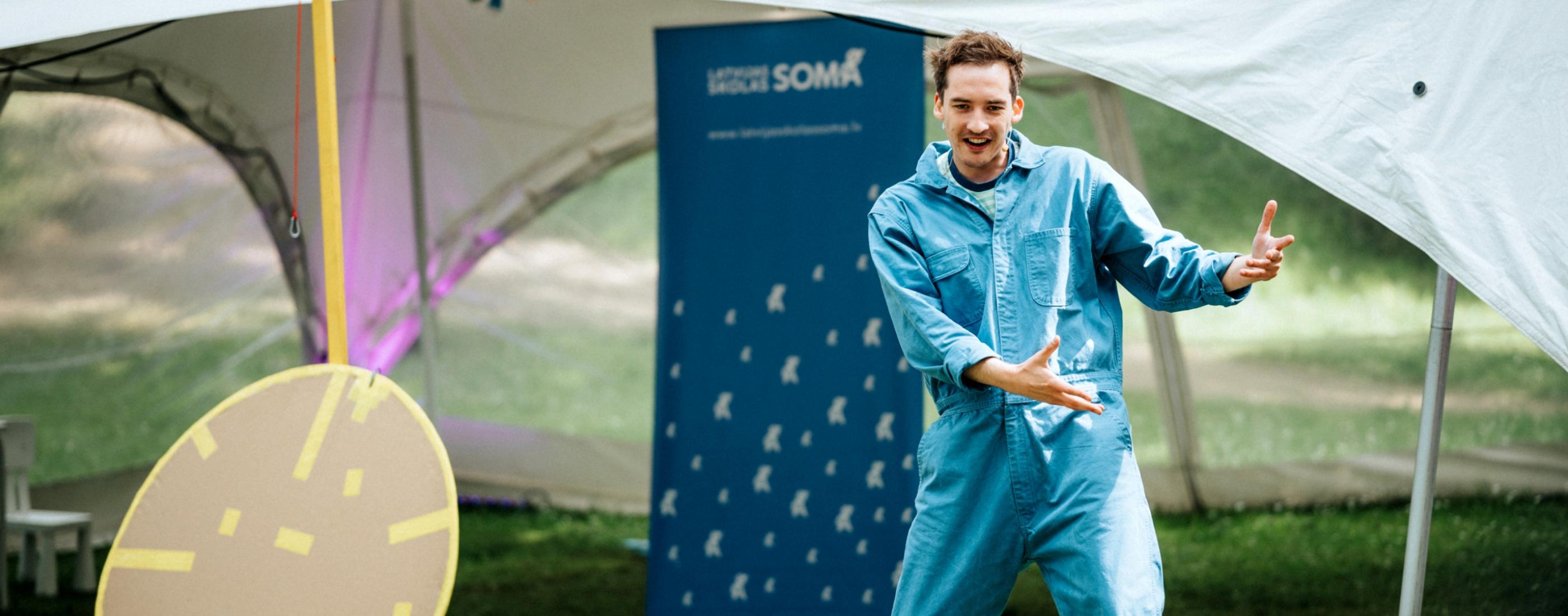 Vīrietis zilā kombinezonā entuziastiski žestikulē āra pasākuma laikā. Zilā banera fonā redzami logotipi, kas rada dzīvīgu atmosfēru.