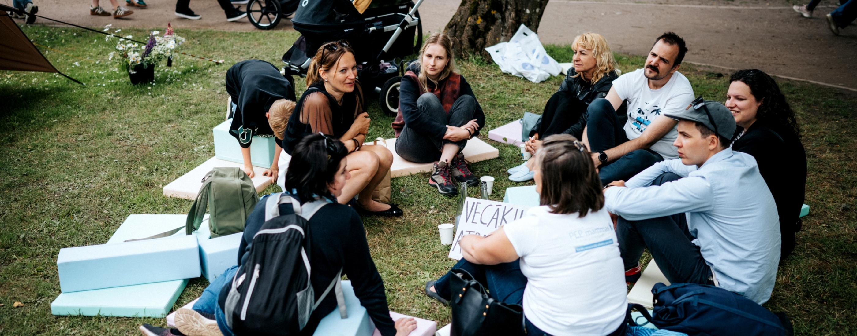 Cilvēku grupa sēž aplī uz putuplasta paklājiņiem zem koka, iesaistījušies sarunā. Netālu redzami ratiņi un papīra zīme.