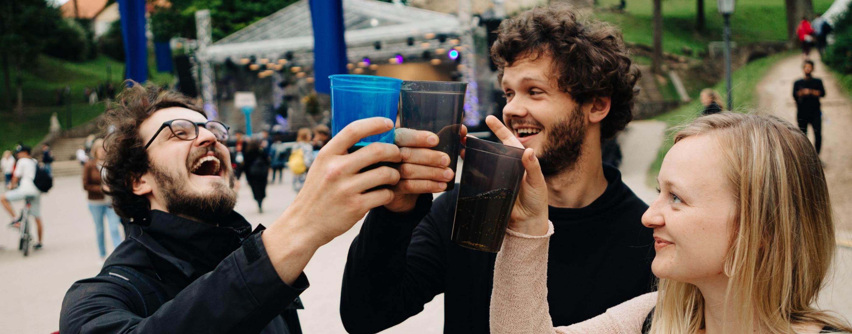 Trīs cilvēki jautri paceļ plastmasas krūzītes brīvdabas pasākumā. Viņi smaida un, šķiet, izbauda rosīgo festivāla atmosfēru.