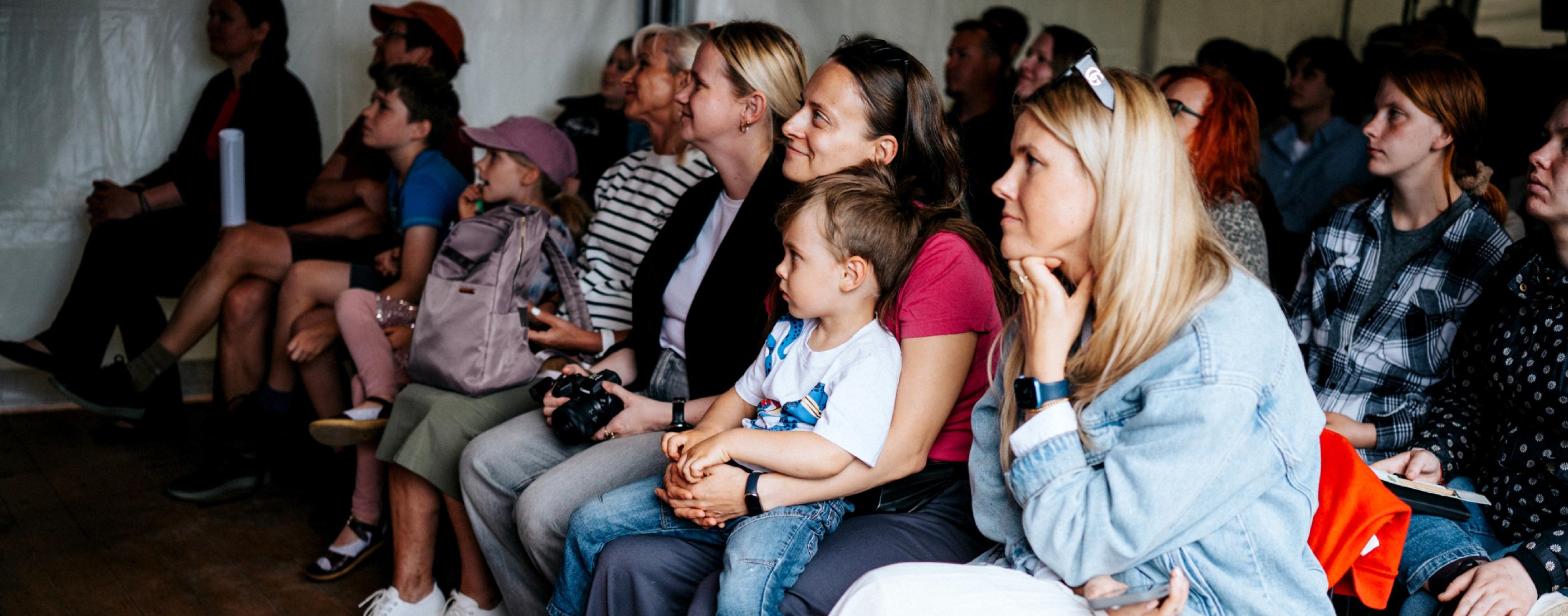 Grupa uzmanīgi sēž, sievietei klēpī sēž bērns, vīriešu un sieviešu ieskauti, radot siltu, aizraujošu atmosfēru. Daži tur rokās kameras.