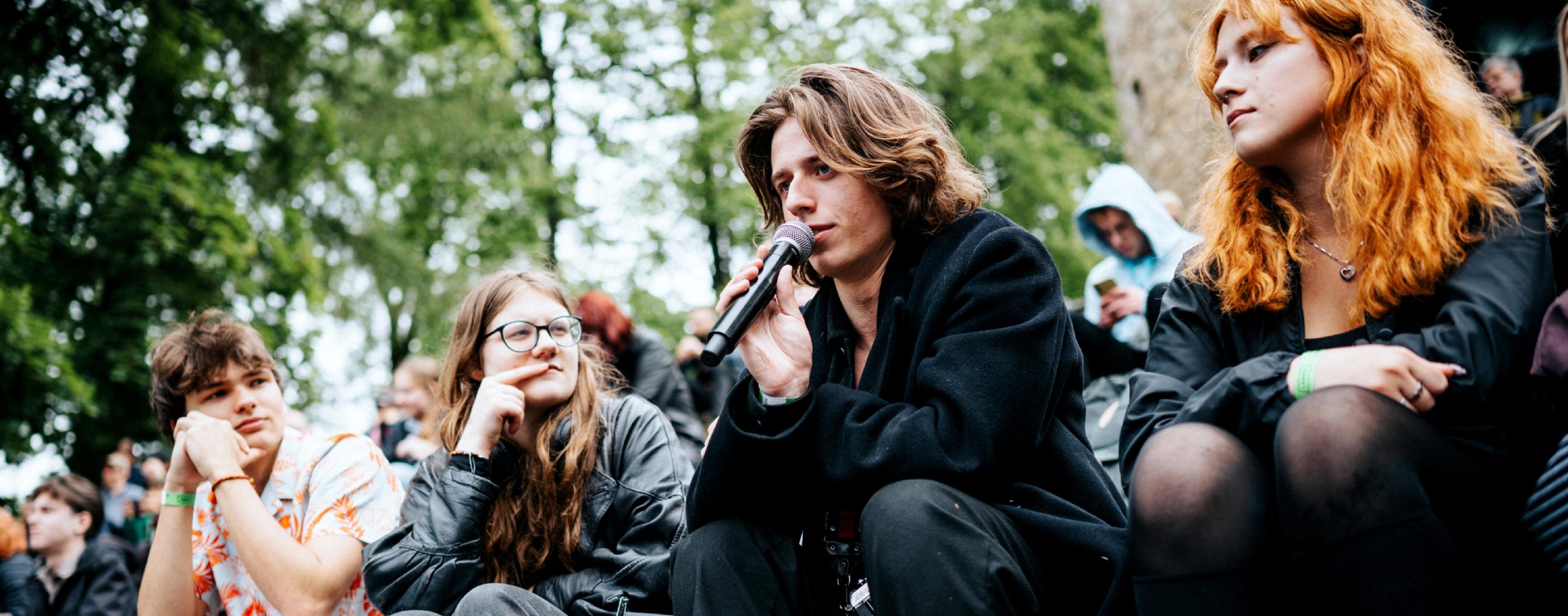 Jauns cilvēks melnā mētelī un zābakos runā mikrofonā, sēžot ārā uz akmens pakāpieniem, uzmanīgiem klausītājiem un kokiem ielenkts.