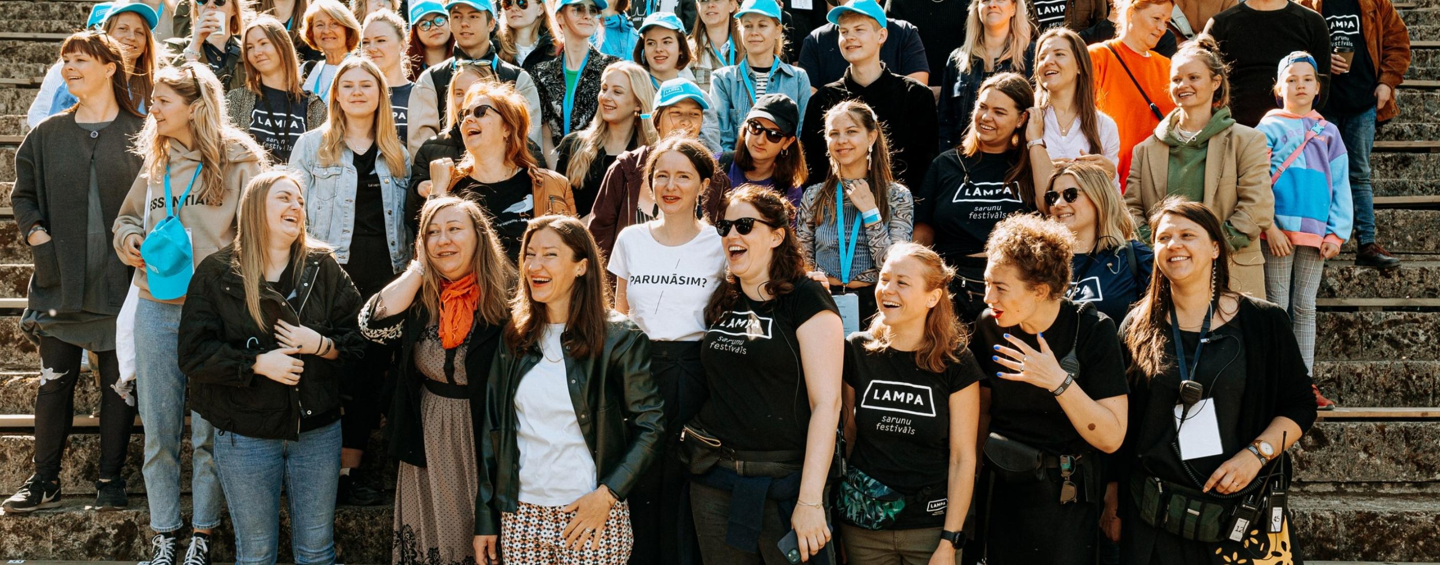 Liela dažādu cilvēku grupa stāv cieši kopā uz pakāpieniem, smaida un smejas. Priekšplānā redzama Sarunu festivāla LAMPA rīkotāju komanda. Fonā redzami festivāla brīvprātīgie ar zilām cepurēm galvās.