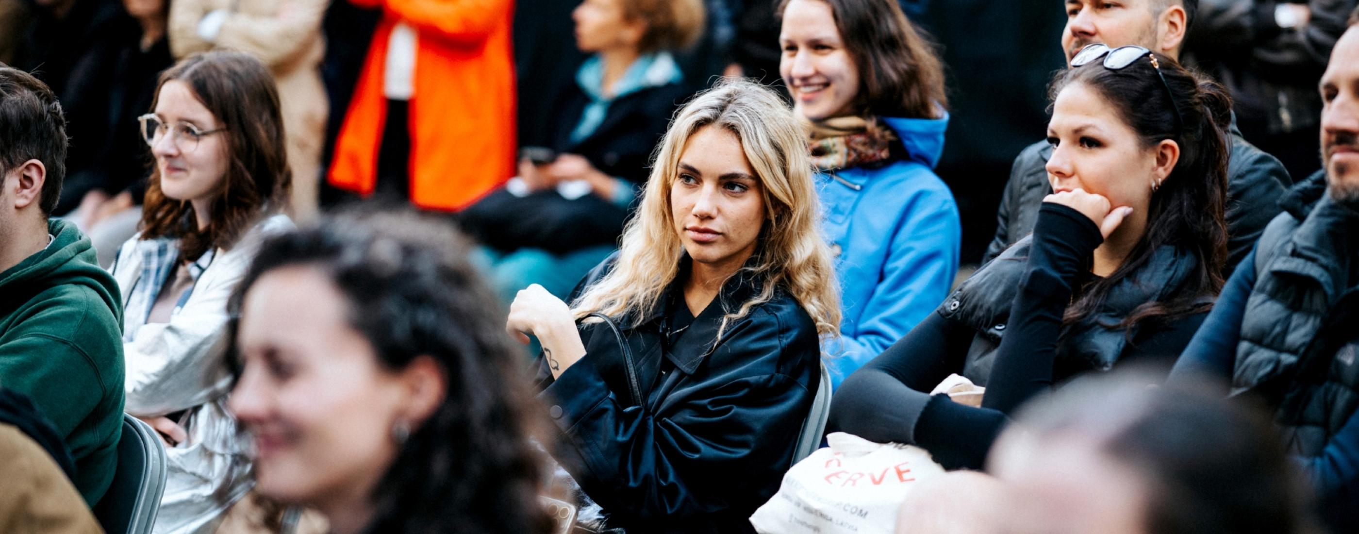 Cilvēku grupa uzmanīgi sēž ārā, ģērbušies vēsam laikam. Atmosfērā valda koncentrēšanās un zinātkāre, daži smaida un ir iesaistīti.