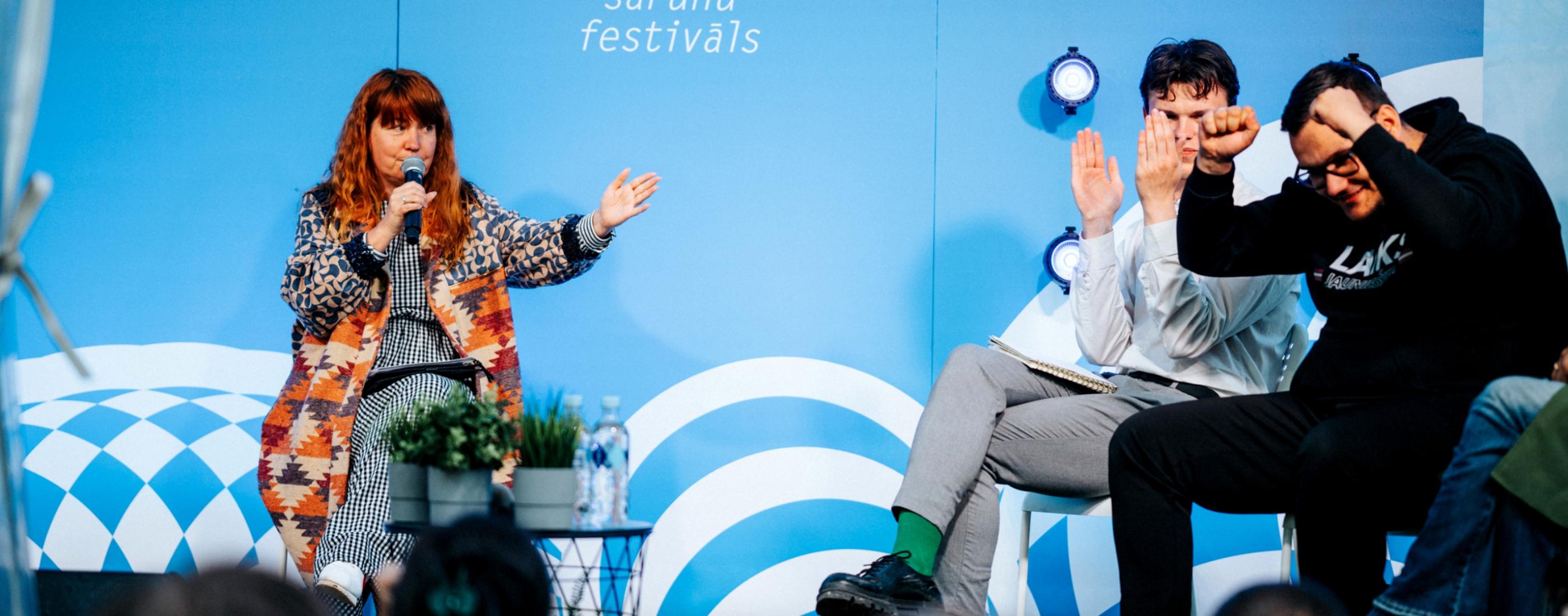 Sieviete ar rudiem matiem dzīvīgi runā mikrofonā Sarunu festivālā LAMPA , sēžot blakus diviem vīriešiem. Fons ir spilgti zils.