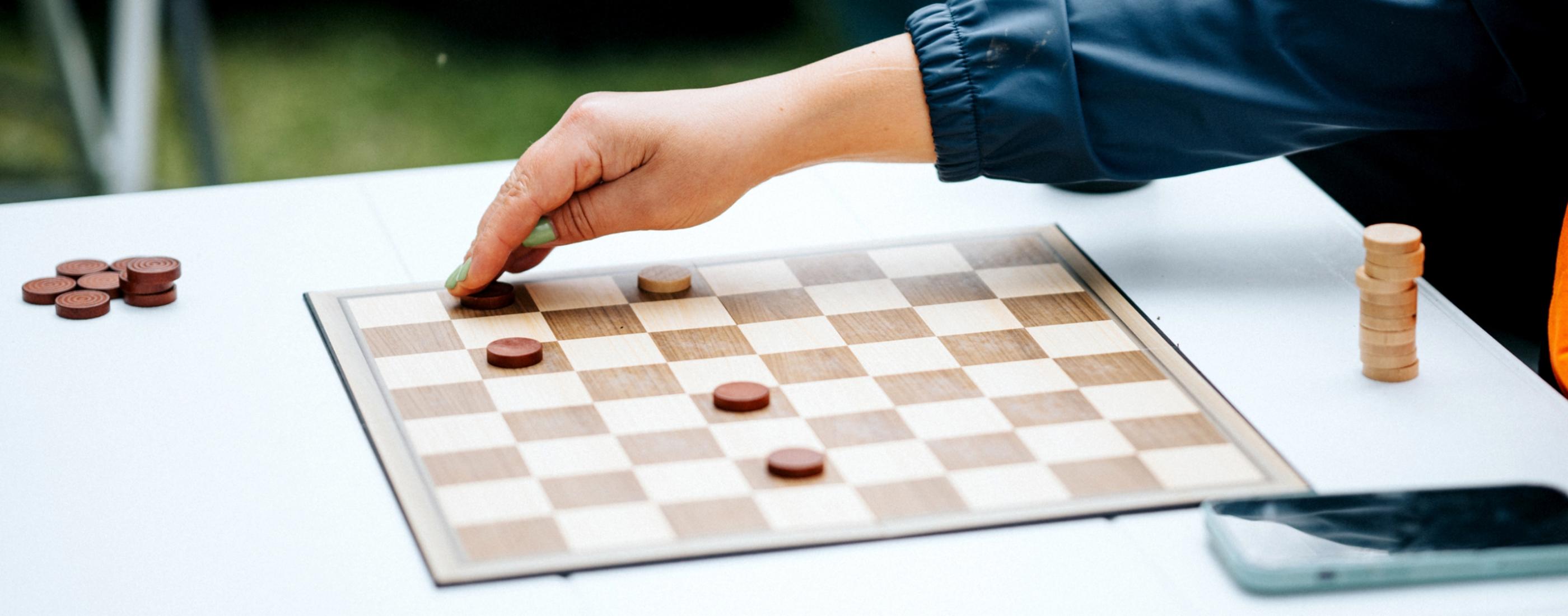 Roka zilā piedurknē pārvieto figūru uz dambretes galdiņa, redzamas piecas figūriņas. Aina atrodas ārā, radot relaksētu, stratēģisku atmosfēru.