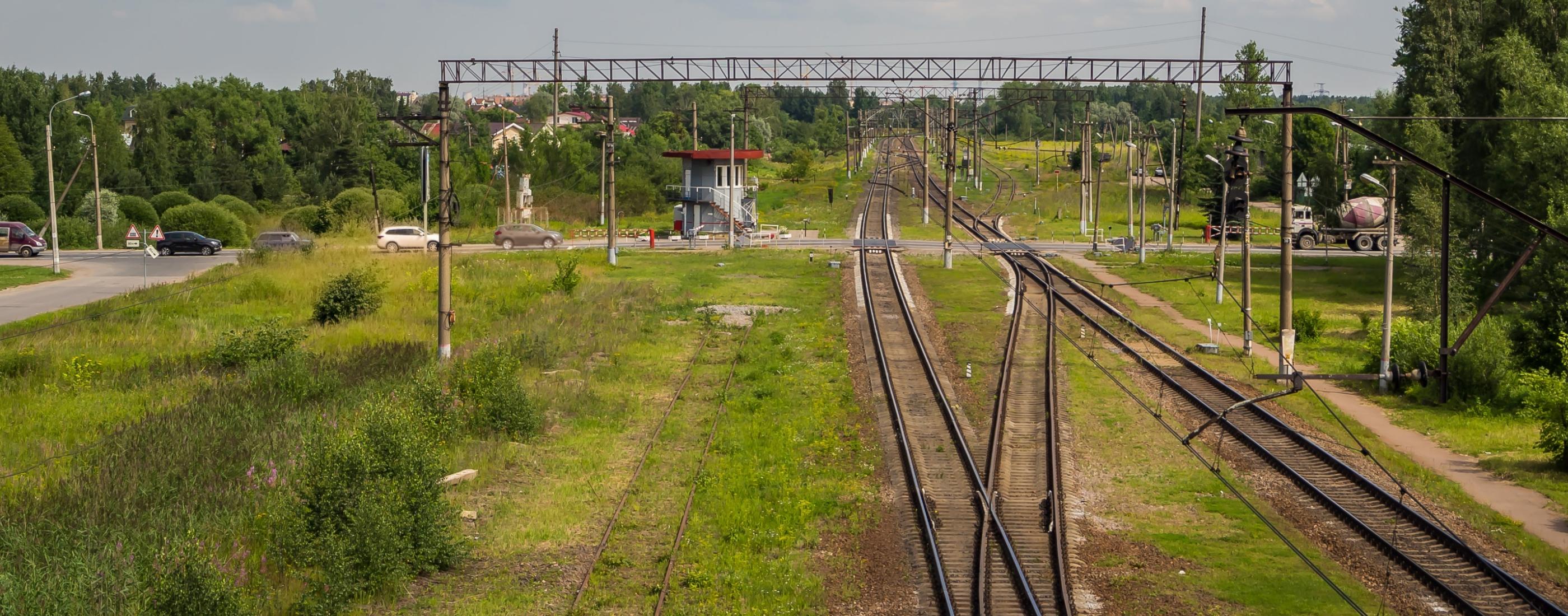 Tālumā stiepjas dzelzceļa sliedes, ko ieskauj zaļi lauki un koki. Kreisajā pusē atrodas neliela signālkabīne ar sarkanu jumtu, un netālu šķērso ceļu automašīna.
