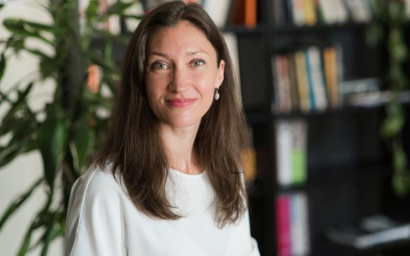 Ieva Morica baltā kreklā maigi smaida. Viņa sēž istabā, kuras fonā ir augs un izplūdis grāmatu plaukts.