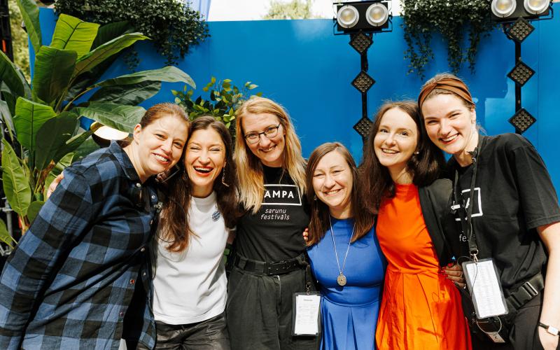 Sešu sieviešu grupa stāv cieši kopā, smaidot uz spilgti zila fona ar zaļiem lapotnēm. Noskaņojums ir priecīgs un svinīgs.