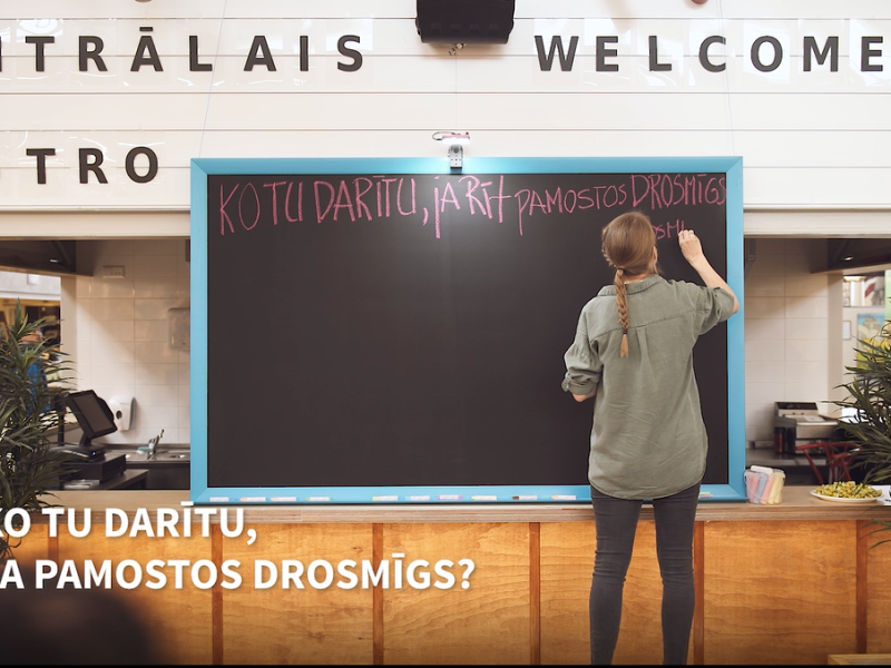 Kafejnīcā cilvēks raksta uz tāfeles.  Priekšplānā teksts: "Kotu darītu, ja pamostos drosmīgs?".Ainu ierāmē augi, radot viesmīlīgu atmosfēru.