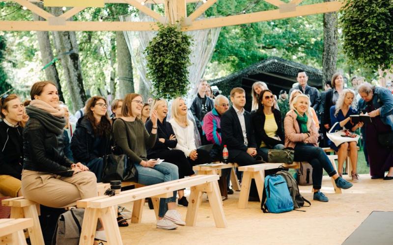 Dažādu cilvēku grupa sēž uz koka soliem zem nojumes, uzmanīgi un ar smaidiem vērojot prezentāciju. Norises vieta ir ārā, koku ieskauta, ar relaksējošu un saistošu atmosfēru. Piekārti augi piešķir ainavai zaļumu.