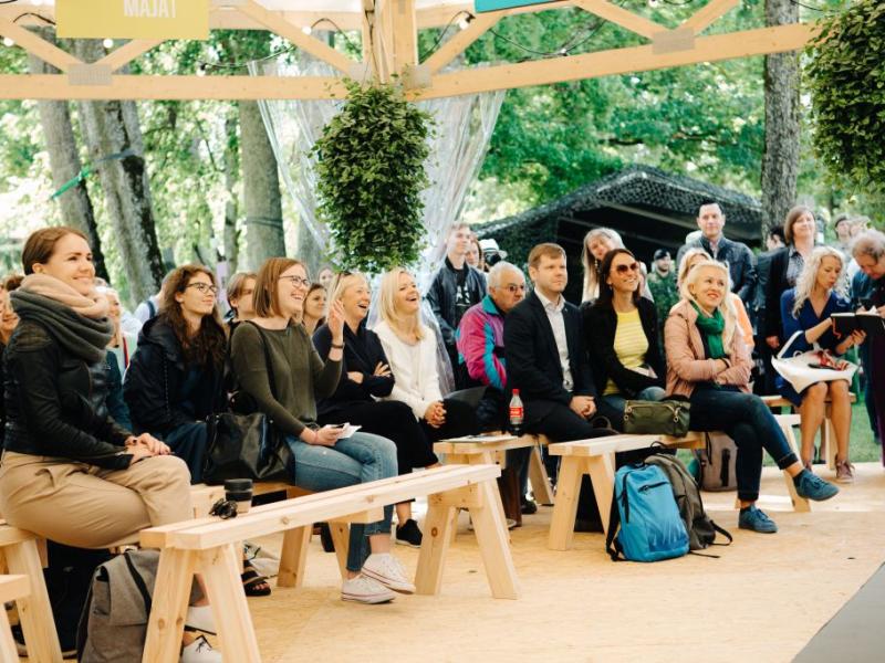 Dažādu cilvēku grupa sēž uz koka soliem zem nojumes, uzmanīgi un ar smaidiem vērojot prezentāciju. Norises vieta ir ārā, koku ieskauta, ar relaksējošu un saistošu atmosfēru. Piekārti augi piešķir ainavai zaļumu.