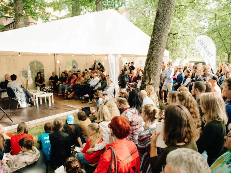 Liela cilvēku grupa uzmanīgi vēro paneļdiskusiju zem baltas telts zaļā parkā. Atmosfēra ir dzīvīga un iesaistīta.