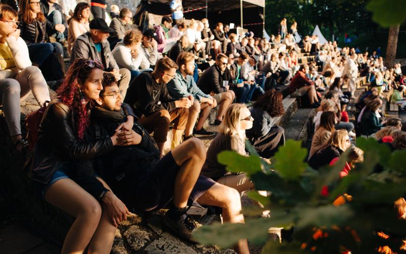 Dažādu cilvēku pūlis sēž uz akmens kāpnēm brīvā dabā, baudot saulainu pasākumu. Atmosfēra ir relaksēta un dzīvespriecīga, un tērpi ir ikdienišķi.
