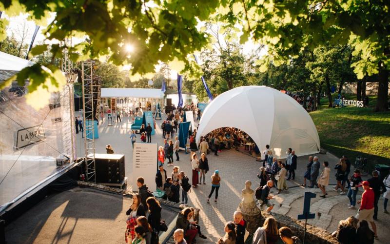 Dinamisks brīvdabas festivāls, kurā pūļi pulcējas ap lielu, baltu telti zem lapu kokiem. Spoža saules gaisma met garas ēnas, radot dzīvīgu un dzīvespriecīgu atmosfēru.