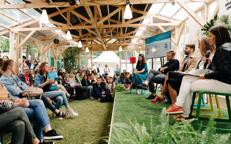 Piecu cilvēku panelis sēž uz skatuves ar mikrofoniem, iesaistot sēdošu auditoriju siltumnīcai līdzīgā struktūrā. Tonis ir dzīvīgs un interaktīvs.