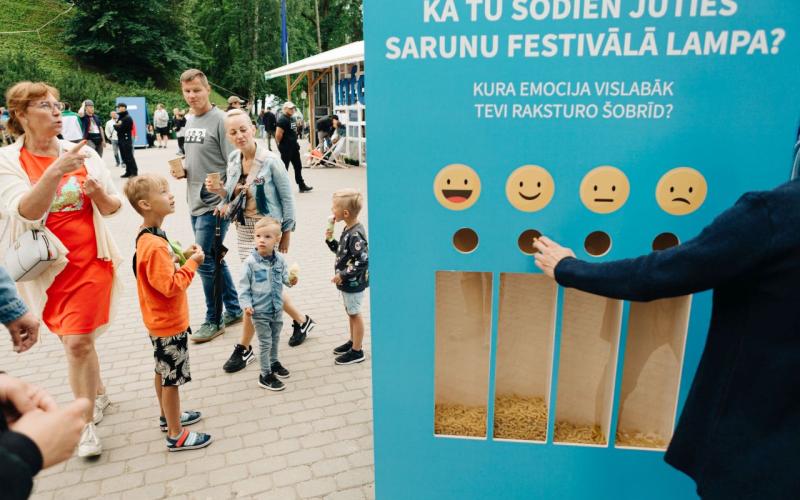 Cilvēku grupa stāv pie tirkīzzilas atsauksmju tāfeles ar dažādiem emocijzīmju simboliem emociju izvēlei.