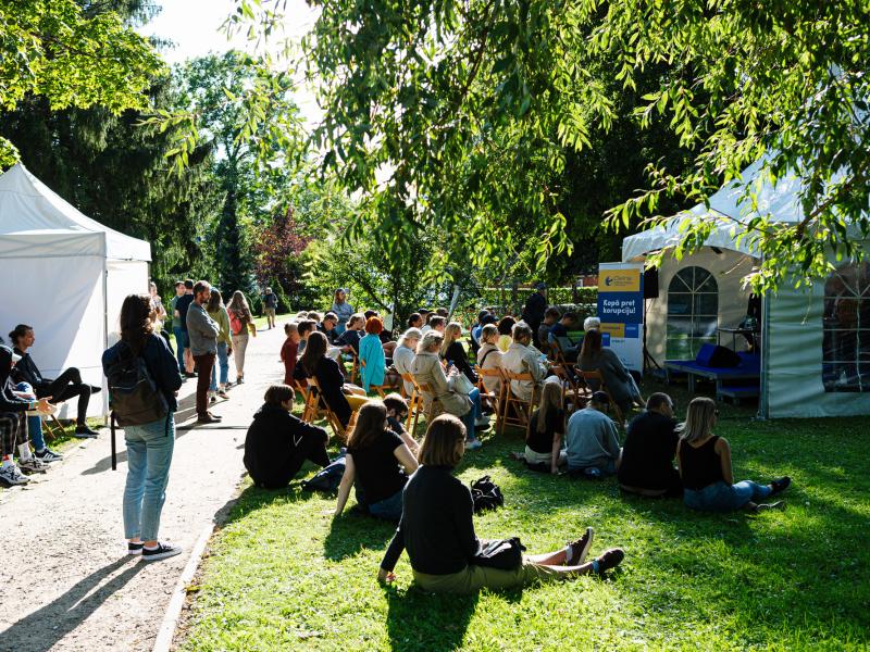 Pulcēšanās brīvā dabā saulainā parkā, kur cilvēki sēž uz krēsliem un zālē, klausoties runātājā pie baltas telts. Koki rada mierīgu fonu.