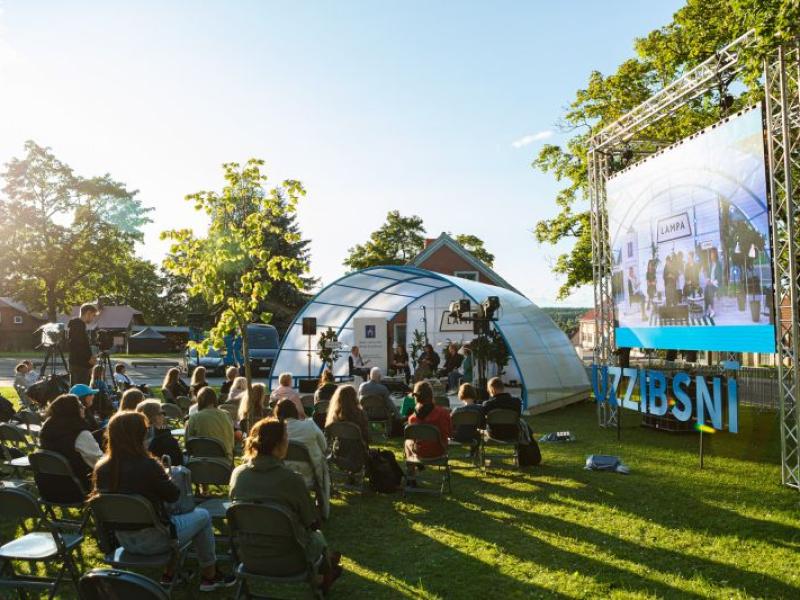 Āra pasākums saulrietā parkā ar sēdošu auditoriju, kas vērsta pret skatuvi kupola teltī. Pasākums tiek rādīts uz liela ekrāna. Apkārtni ieskauj koki.