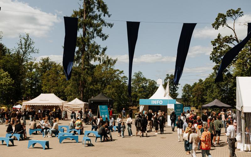 Āra pasākums, kurā cilvēki pulcējas ap teltīm ar uzrakstu "INFO". Zili soliņi un karogi rada dzīvīgu, svētku atmosfēru zem skaidrām debesīm.